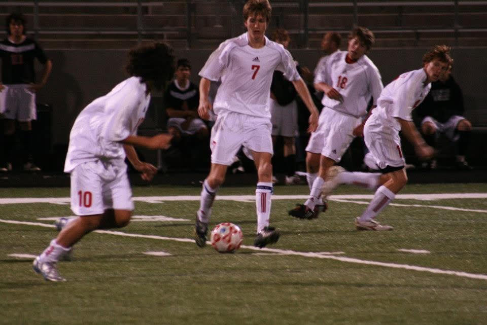 Dr. Stephens on the ball in midfield during a night soccer match, teammates and opponents converging